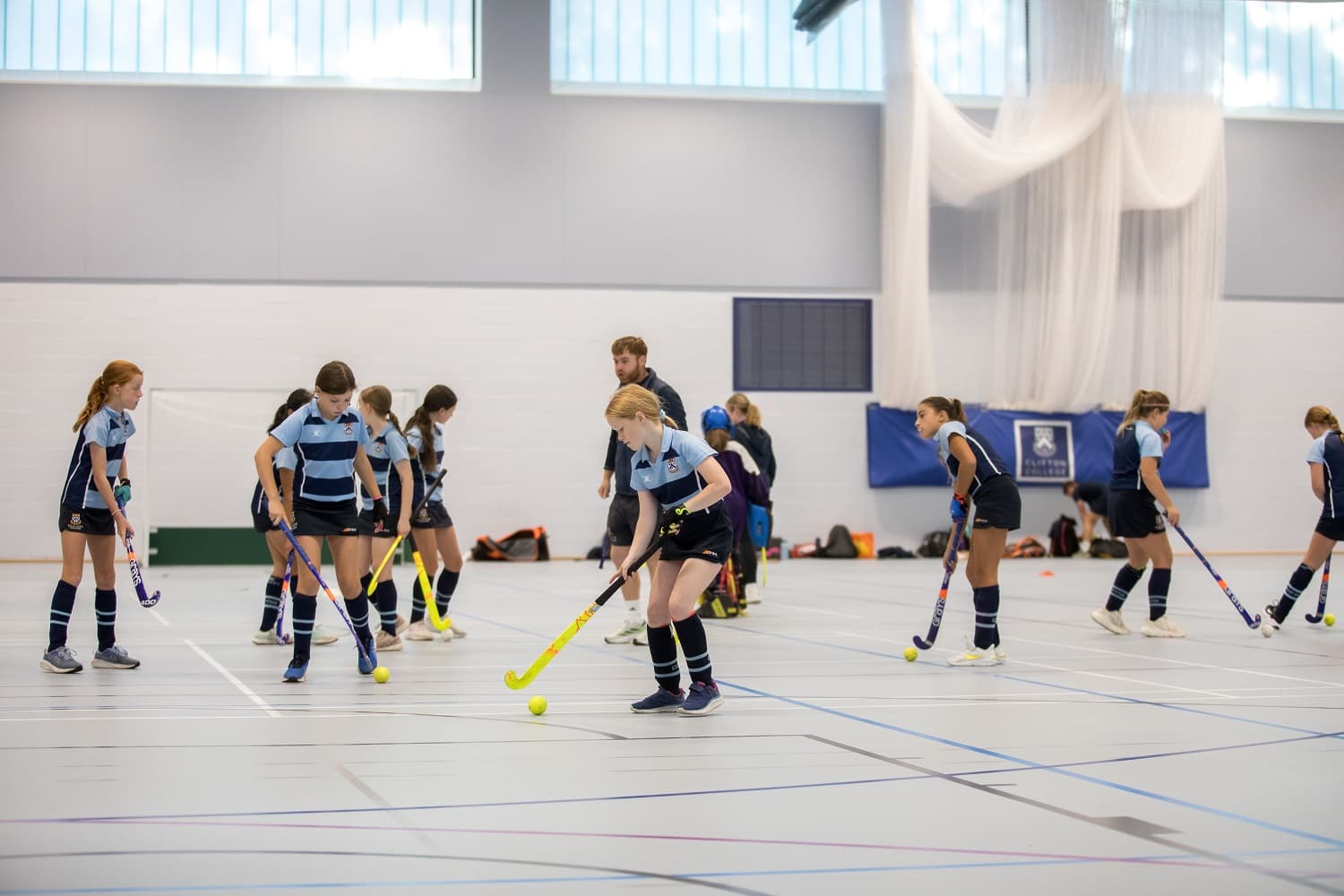 A group of young players in blue uniforms practice field hockey indoors, focusing on ball control with sticks on a spacious court.