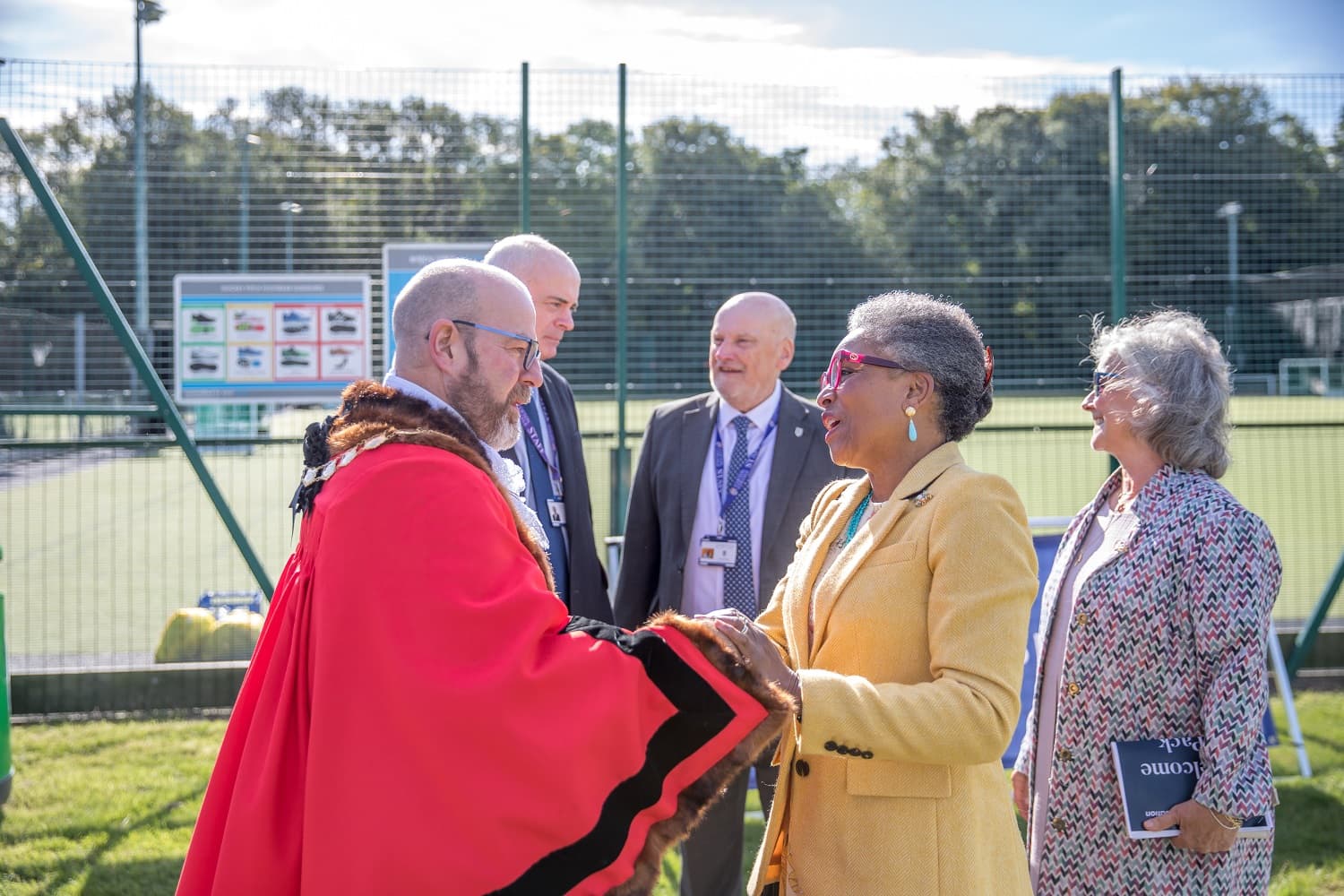 A man in a red robe greets a woman in a yellow jacket outdoors, with three people standing nearby on a sunny day near a sports field.