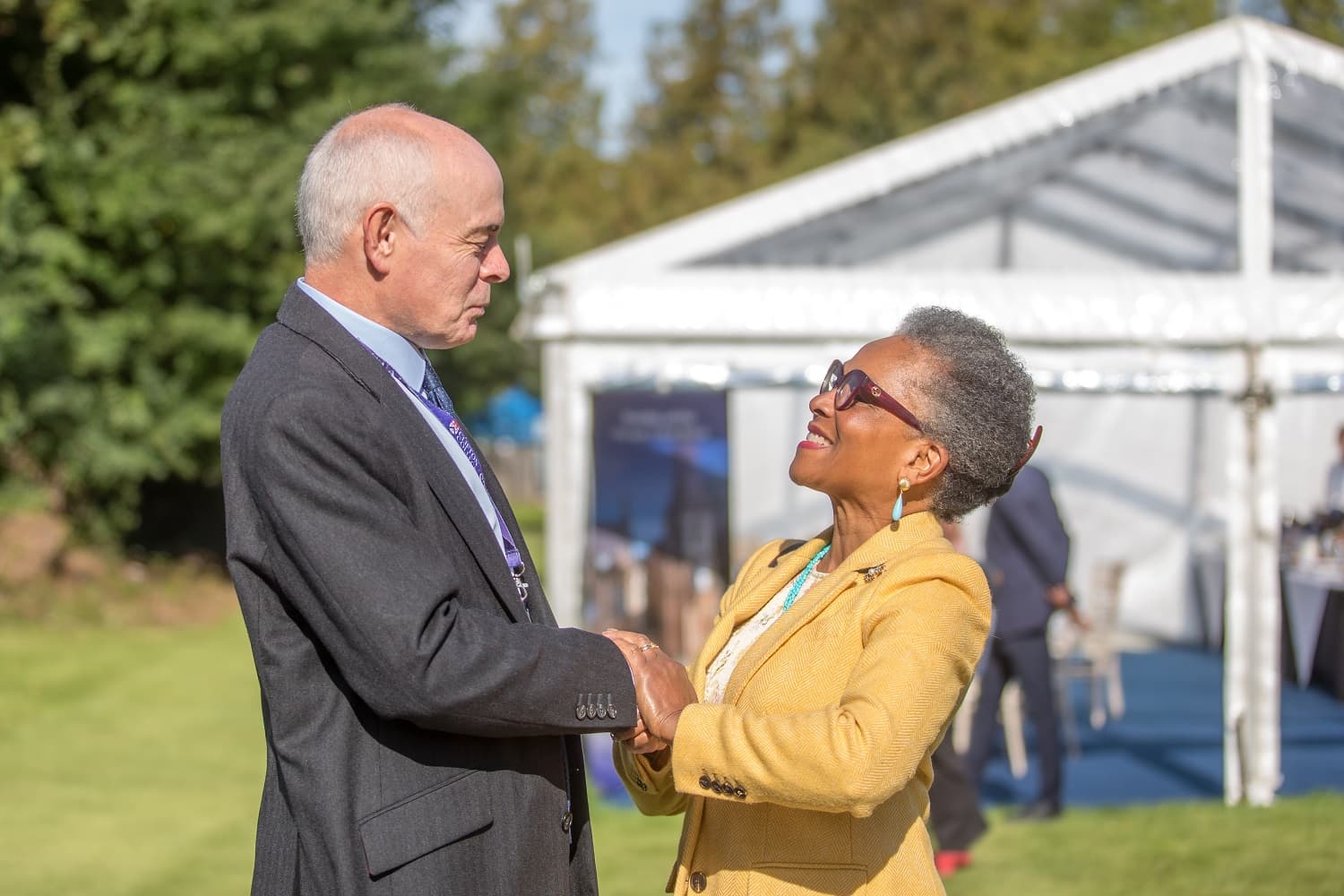 An elderly man and woman smiling and shaking hands outdoors near a white tent on a sunny day.