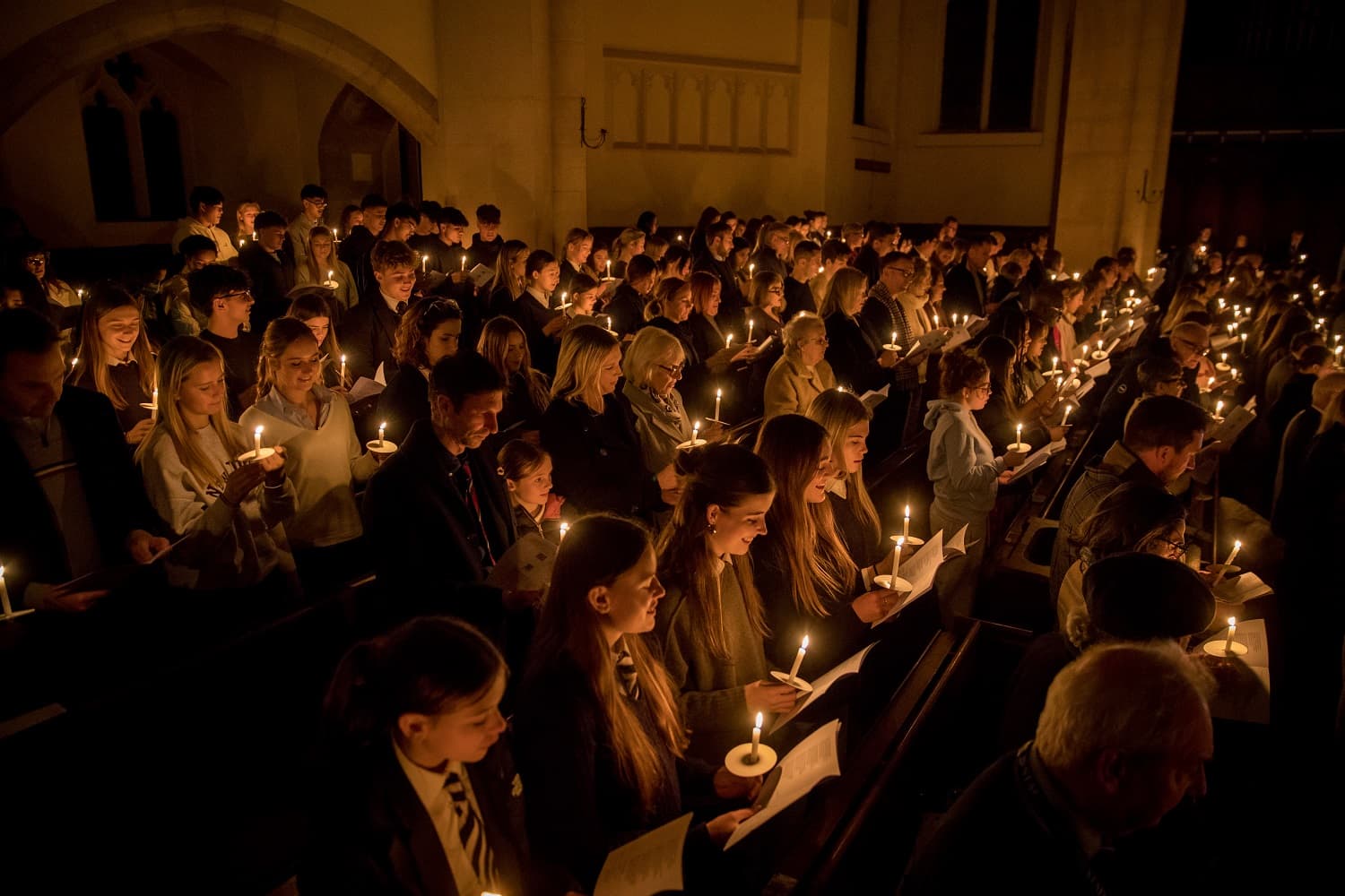 A large group of people in a dimly lit chapel hold candles, creating a warm glow. They appear to be participating in a solemn ceremony.