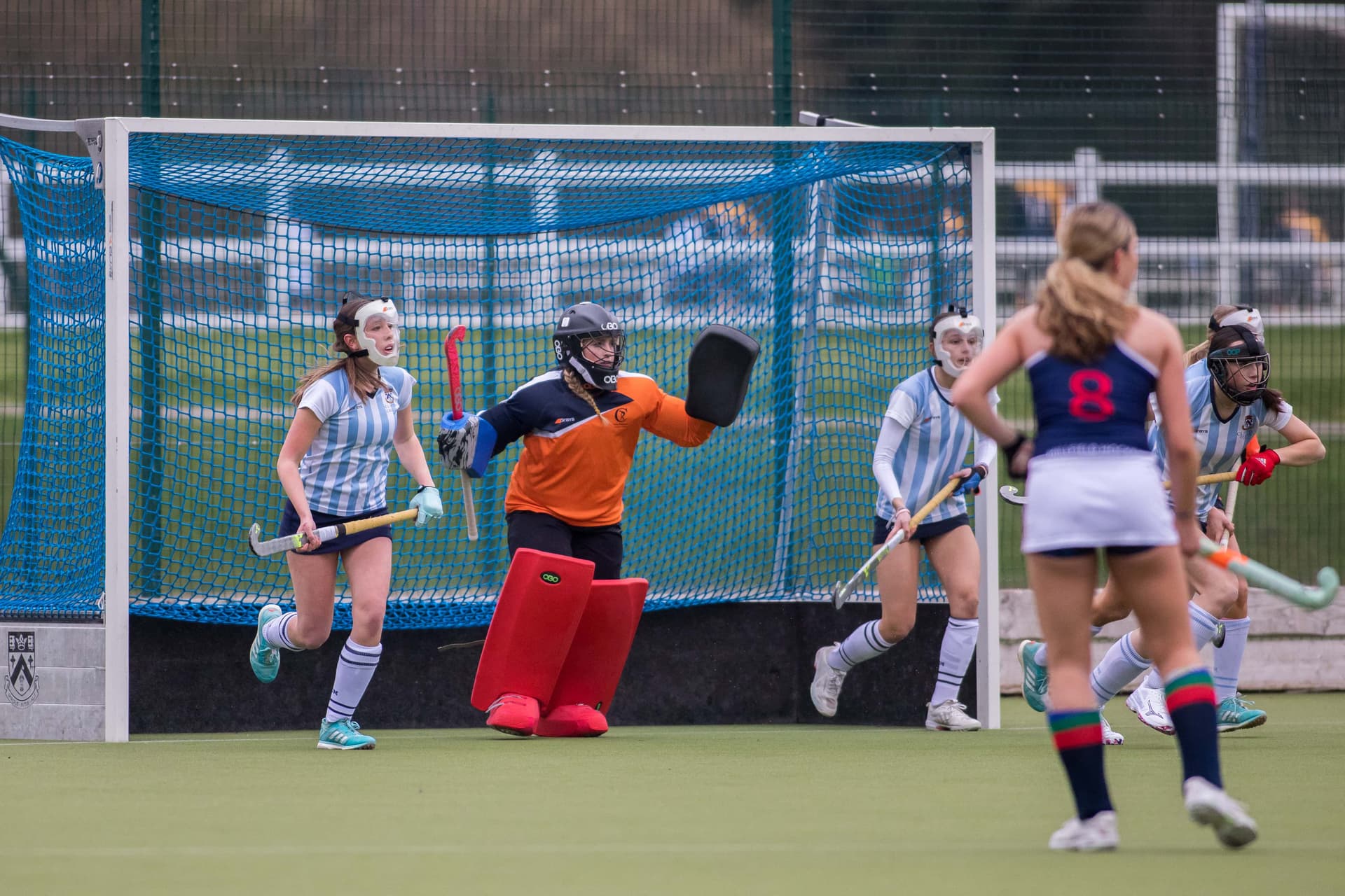 Field hockey game in action, with a goalie in orange and red gear defending the goal as players in blue and white stripes approach.