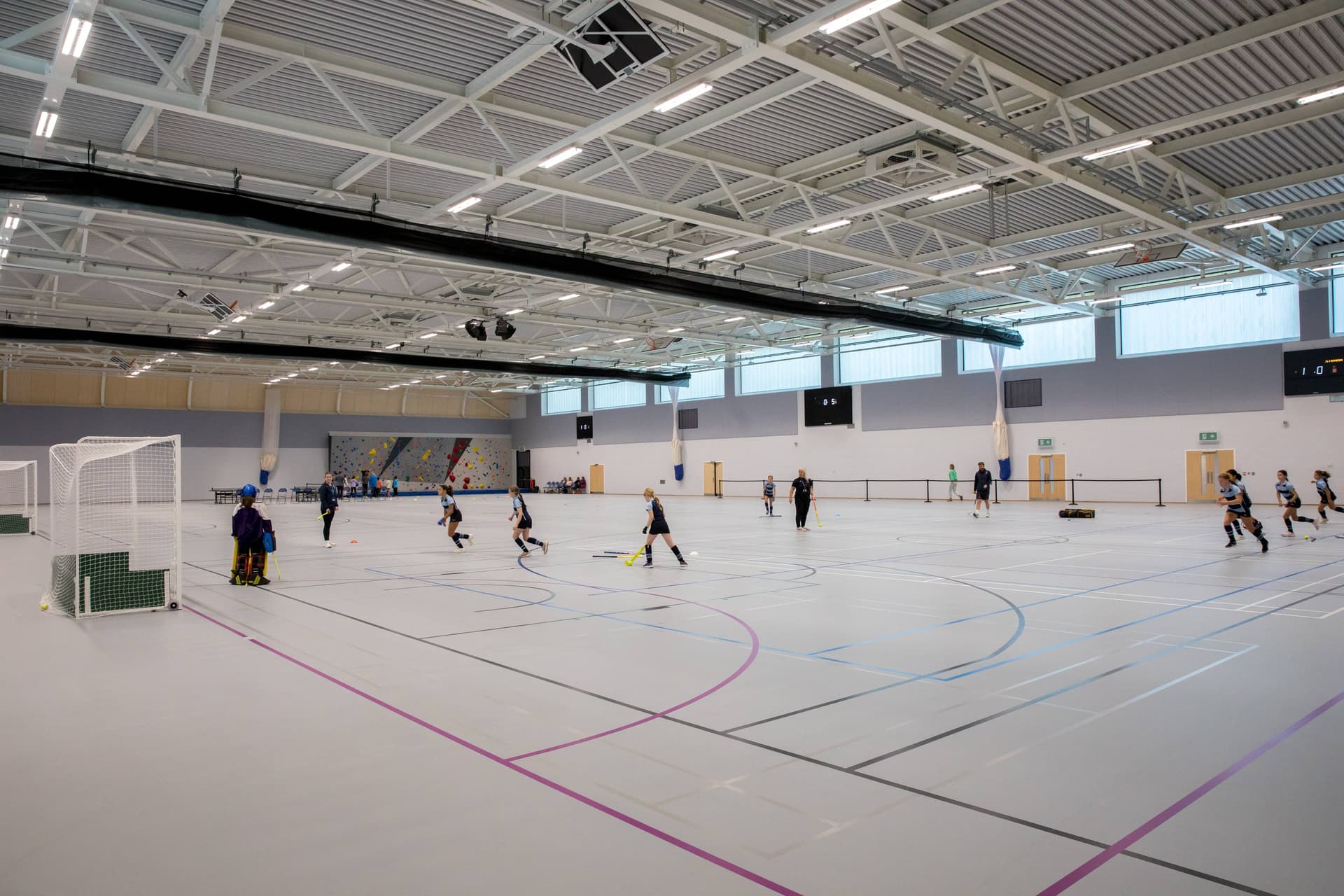 Indoor sports hall with players practicing field hockey. The space has a high ceiling, large windows, and a climbing wall in the background.