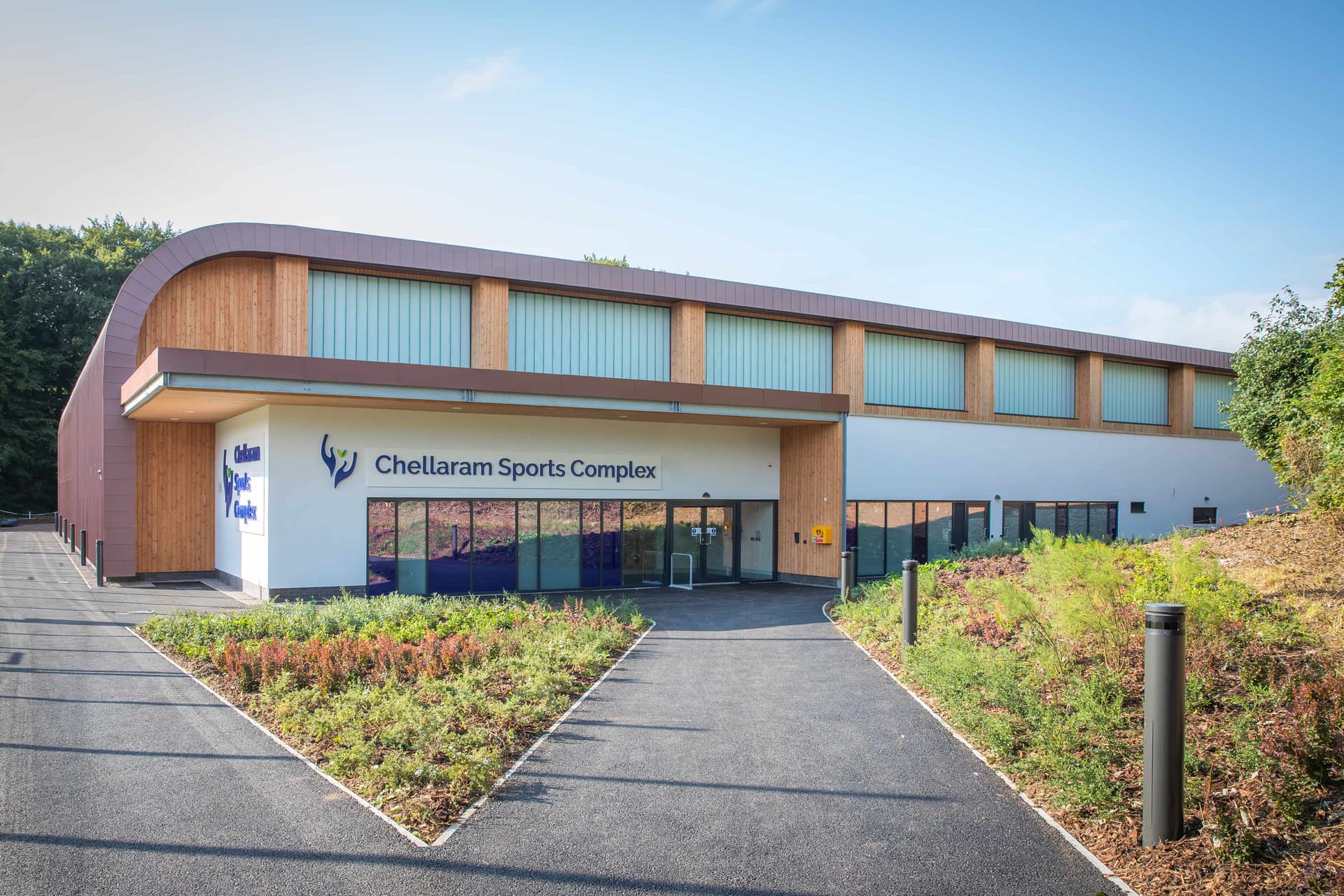 Modern sports complex with large windows, wooden accents, and "Chellaram Sports Complex" signage, surrounded by landscaped greenery.
